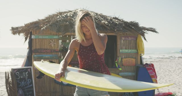 Blond woman standing near a rustic surf shop on sandy beach, getting her surfboard ready. Sunlight shines brightly, creating a summer vibe. This scene is ideal for promoting beachwear, surfing equipment, summer holiday destinations, or relaxation activities.