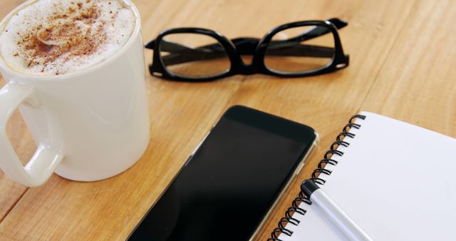 Workspace with a cup of cappuccino, a smartphone, black-rimmed glasses, and a spiral notepad with a pen on a light wooden desk. Ideal for promoting productivity, work-from-home setups, lifestyle blogs, office supplies, or digital planning tools.