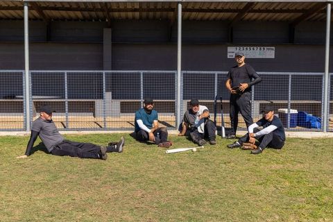 Diverse baseball team relaxing at dugout sitting on grass holding bats and gloves