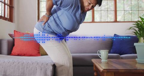 Elder indian woman rising from sofa holding lower back wincing from hip pain at home