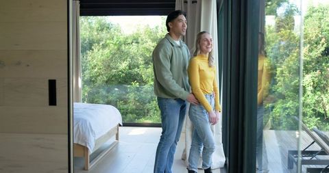 Multicultural couple embracing by glass doors overlooking sunlit garden from modern bedroom