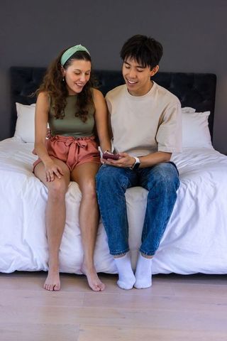 Diverse couple sitting on bed sharing smartphone, relaxing in cozy modern bedroom