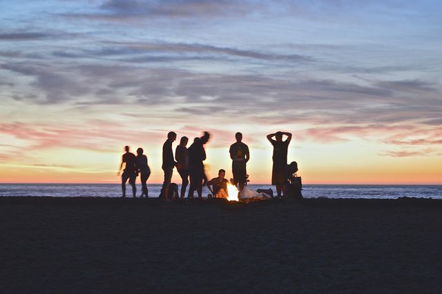 Group of friends standing and sitting around a bonfire on beach during sunset. Vibrant colors of sky illuminate ocean in background, creating warm and inviting atmosphere. Perfect for themes related to summer activities, friendship, relaxation, nature, and vacations. Ideal for promoting travel destinations, outdoor products, and lifestyle content.