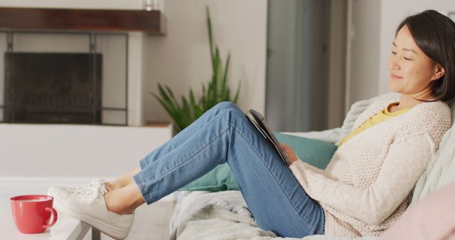 Woman enjoying a moment of relaxation on a sofa, reading content on a tablet in a comfortable home setting. Perfect for articles or promotions regarding leisure, home lifestyle, technology usage, and cozy living spaces. Captures the essence of a peaceful indoor environment and modern living.