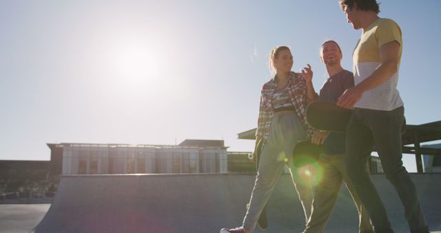 Group of young friends walking together at skate park while holding skateboards. Smiling and enjoying conversation in casual clothing under bright sunlight. Ideal for social media, friendship, outdoor activities, youth culture and lifestyle concepts.