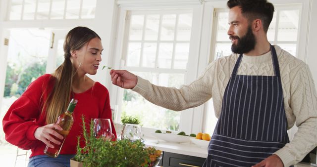 Couple enjoying cooking together in a modern kitchen. The man in an apron feeds a woman holding a bottle of wine. They are smiling and enjoying each other's company. Ideal for concepts related to love, relationships, home life, cooking, and lifestyle blogs.