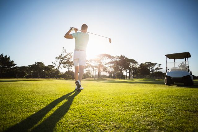 Male golfer taking a shot on a sunny golf course with a golf cart nearby. Ideal for use in sports and recreation advertisements, golf-related promotions, outdoor activity campaigns, and leisure lifestyle content.