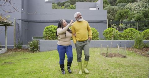 Senior couple walking arm-in-arm across backyard lawn pointing toward garden in boots