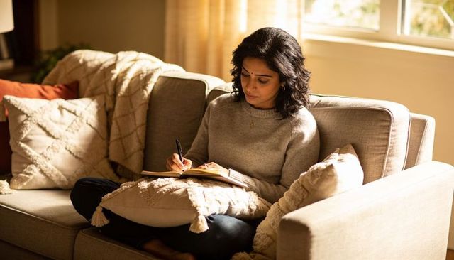 Sunlit south asian woman journaling on cozy beige sofa wearing grey knit sweater and jeans