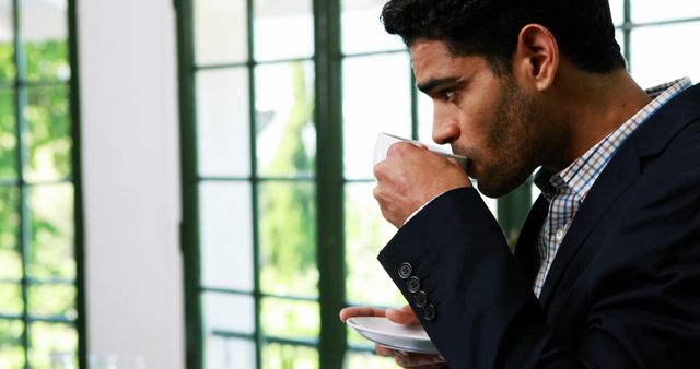 A middle-aged man in a business suit enjoys a cup of coffee by a window, with copy space. His moment of reflection or break from work illustrates the importance of taking short breaks to recharge during a busy day.