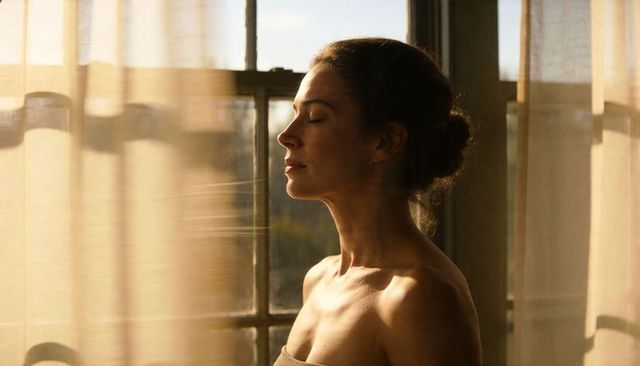 Woman standing by sunlit window breathing with eyes closed for morning calm mindfulness