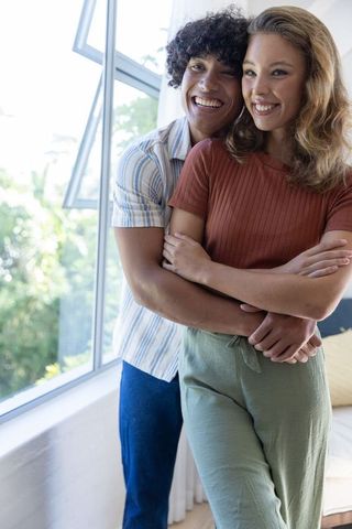 Young couple hugging by sunlit casement window in cozy home, smiling partners and affection