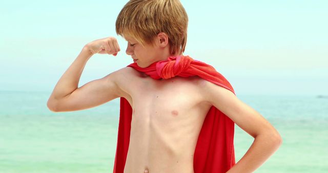 Young blond boy in red cape flexing muscles by beach - Download Free Stock Images Pikwizard.com