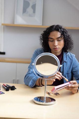 Non-binary adult applying makeup at home with mirror and pink palette on wooden table