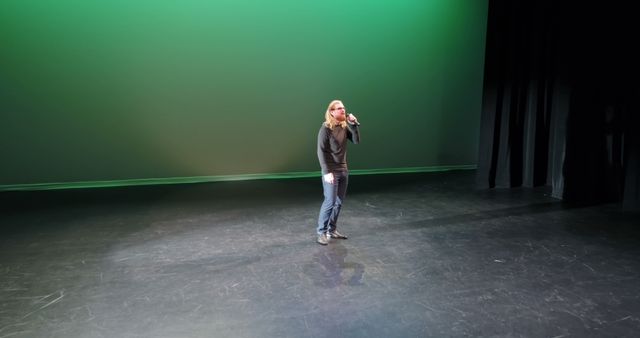 A Caucasian man stands alone on a stage with a green background, holding a microphone, with copy space. His casual attire and solitary presence suggest a rehearsal or sound check in a performance setting.