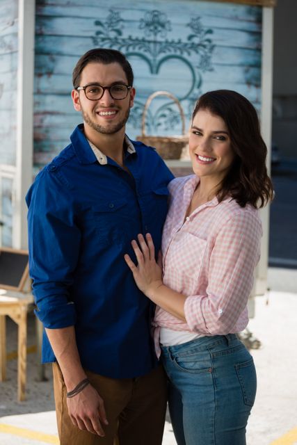 This image captures a happy couple standing close together and smiling at an outdoor cafe. They are dressed in casual clothing, with the man wearing a blue shirt and glasses, and the woman in a pink shirt and jeans. This image is perfect for use in advertisements, relationship blogs, lifestyle articles, and social media posts promoting love, togetherness, and casual urban living.