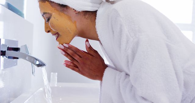 Young woman wearing white bathrobe and towel, cleansing face with homemade orange face mask, standing in modern bathroom. Perfect for articles on natural skincare routines, beauty tutorials, spa day inspiration, self-care practices, and wellness blogs.