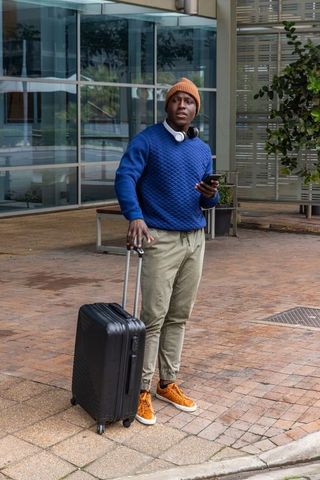 African american man waiting with suitcase and smartphone outside modern office building