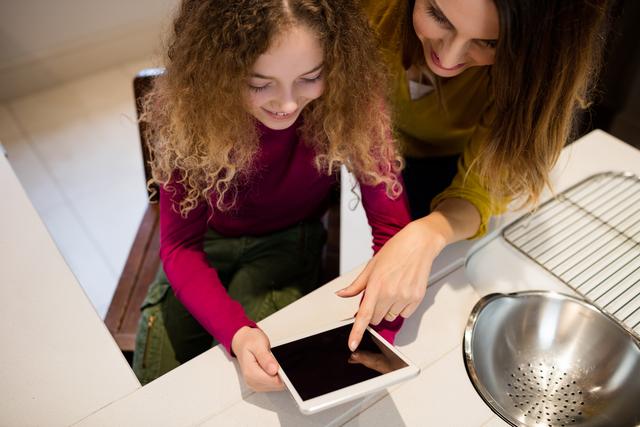 Mother and daughter engaging with a digital tablet in a modern home kitchen. They are smiling and enjoying their time together, which highlights family bonding, learning, and the use of technology at home. This can be used for themes related to parenting, family time, education, modern lifestyle, and home activities.
