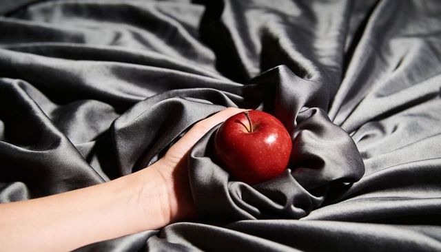 Hand cradling glossy red apple on draped gray satin fabric, dramatic lighting