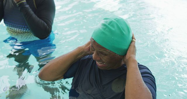 Senior woman is enjoying a relaxing time in a swimming pool wearing a green swimming cap and dark swimwear. She is smiling joyfully, suggesting a moment of leisure and contentment. Another person is partially visible in the background, also in the water. This image can be used for promoting senior health, retirement activities, leisure and wellness programs targeted at older adults, or aquatic exercise routines.