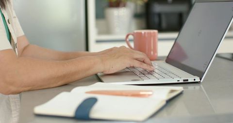 Senior woman typing on slim laptop at kitchen countertop, home office productivity scene