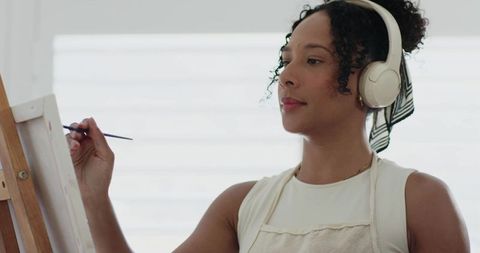 African american woman painting on canvas wearing headphones and apron in bright studio