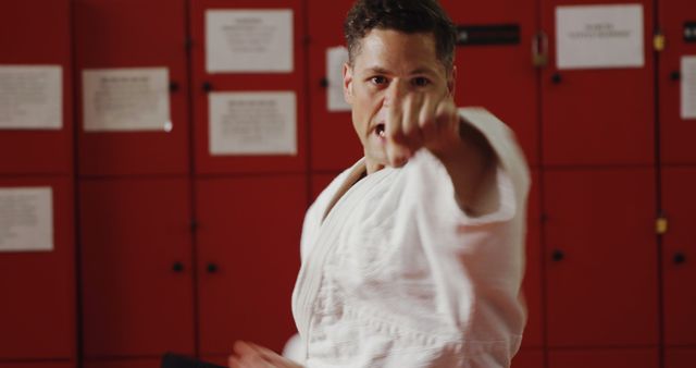 Man practicing a karate punch in a locker room, wearing white gi, focused expression. Highlights martial arts discipline and physical fitness. Useful for websites or promotional materials related to karate, self-defense, fitness training, sportswear, or motivational content.