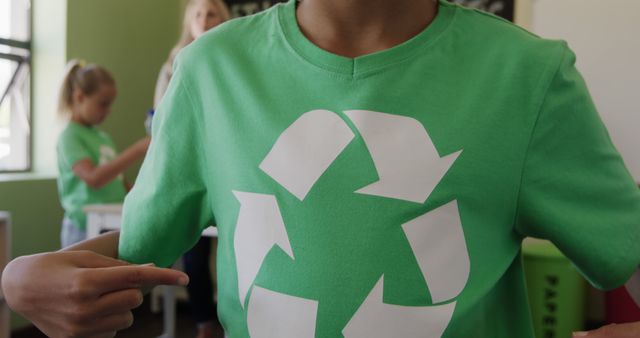 Brightly lit classroom featuring young student pointing to large recycling symbol on green t-shirt. Ideal for promoting environmental campaigns, school sustainability projects, and recycling programs. Illustrates youth involvement in eco-friendly activities and raising awareness about environmental conservation.