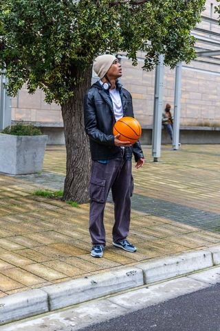 Diverse young man standing holding basketball wearing beanie and headphones in urban plaza