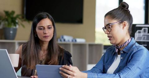 Diverse female coworkers reviewing tablet and collaborating at modern office workspace