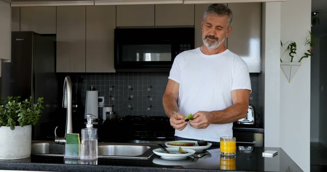 An older man in casual attire preparing breakfast in a modern kitchen. He is slicing an avocado on the kitchen counter, where a glass of orange juice and other kitchen items are placed. This image is ideal for use in lifestyle blogs, health and wellness articles, kitchen appliance advertisements, or senior living guides.