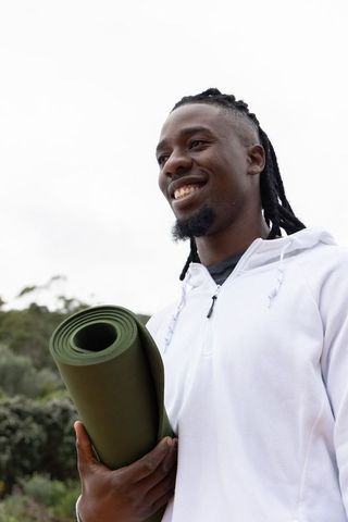 African american man smiling holding olive green yoga mat outdoors in white hoodie