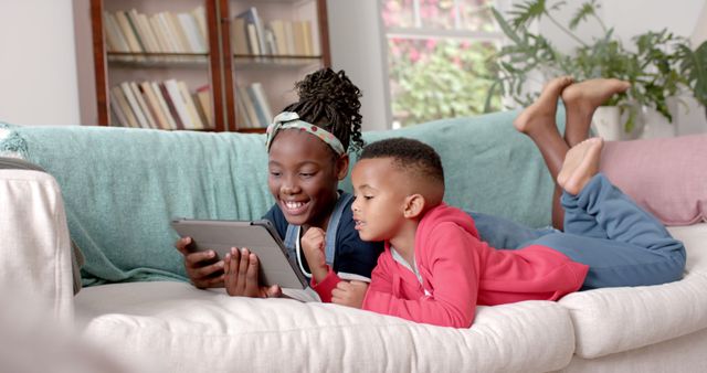 Image shows two happy siblings, a girl and a boy, lying on a couch and using a tablet at home. Ideal for content related to family bonding, children's technology use, digital education, relaxed home environments, and leisure activities. Useful for blogs, educational articles, tech reviews, family lifestyle promotions, and home interior websites.