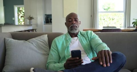 African american senior man relaxing on sofa using smartphone in bright modern living room