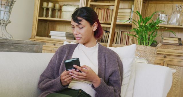 Woman seated on casual couch in cozy living room checking phone, giving impression of relaxed, everyday home setting. Ideal for stock where someone is engaging with or taking a break from technology, personal downtime, or home décor advertisements.
