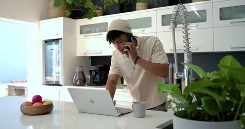 African american man juggling phone call and laptop while working on bright kitchen island