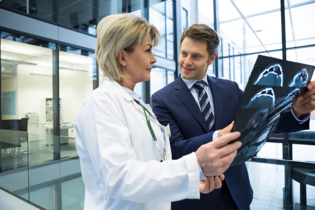 Female doctor in white coat discussing x-ray report with businessman in suit in modern hospital. Ideal for use in healthcare, medical consultation, professional collaboration, and business-related content.