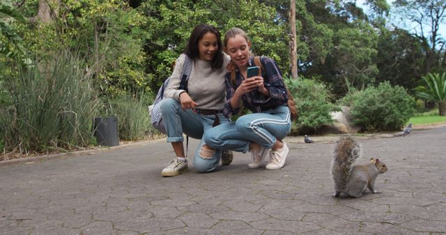 Two friends are eagerly photographing a squirrel in a park. The two young women seem excited while sitting on the pathway surrounded by lush greenery. This image is showcasing leisure activities, is easy, and relaxed, portraying friendship and bonding in a natural outdoor environment. Useful for illustrating themes of youth, nature appreciation, lifestyle content, and outdoor activities.
