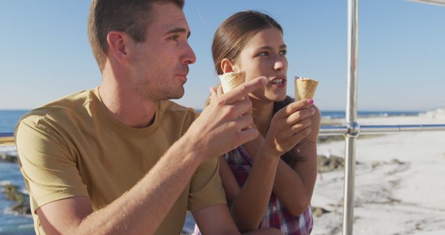 Couple enjoying ice cream cones on beach during sunny day, ideal for summer vacation themes, outdoor relaxation, leisure activities, and travel promotions. Suitable for use in advertisements, travel brochures, and lifestyle blogs.