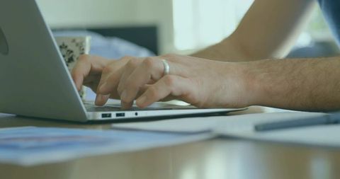 Mature man typing on laptop at home desk with coffee mug, paperwork and wedding ring
