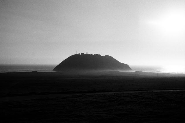Silhouette of a rocky hill against the bright light of the setting sun over the ocean. The serene and scenic view captures the dramatic sky and light, ideal for use in nature and landscape themed projects, travel magazines, environmental campaigns, and wall art showcasing natural beauty and tranquility.