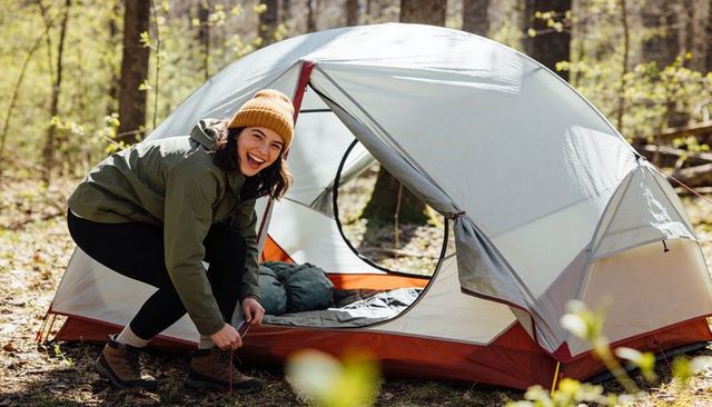 Smiling woman securing tent stake while camping in sunlit forest wearing orange beanie
