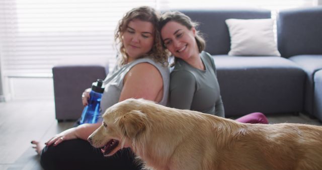 Two women sitting together in living room, smiling and relaxed, with a golden retriever in the foreground. One woman holds a water bottle. Perfect for themes related to friendship, happiness, pet companionship, and leisure activities at home.