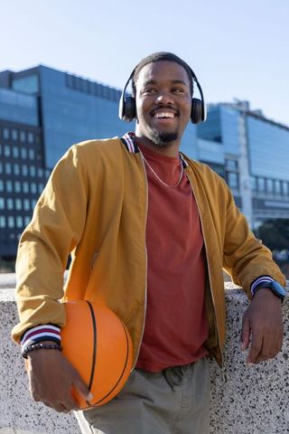African american man holding basketball, wearing headphones, smiling on urban waterfront