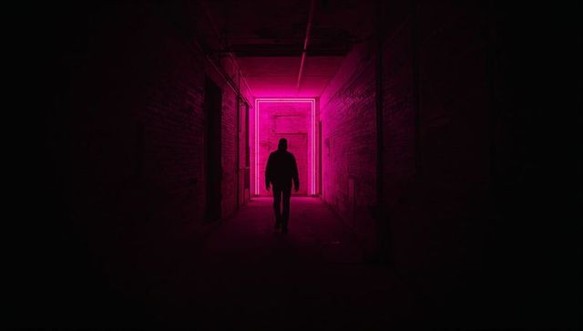 Hooded silhouette walking through neon-lit industrial corridor with magenta light frame
