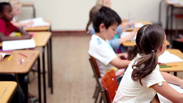 Children are shown intently working at their desks in a classroom featuring wooden furniture. The students seem keen on their tasks, indicating an attentive educational environment. Use this image for educational websites, school brochures, or articles emphasising the importance of focused learning in school systems.