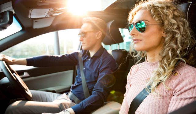 Couple driving car during test drive on sunny day, both wearing sunglasses and seatbelts. Ideal for use in advertisements for car dealerships, automotive safety campaigns, travel blogs, and lifestyle articles. Highlights themes of modern transportation, safety, and adventure.