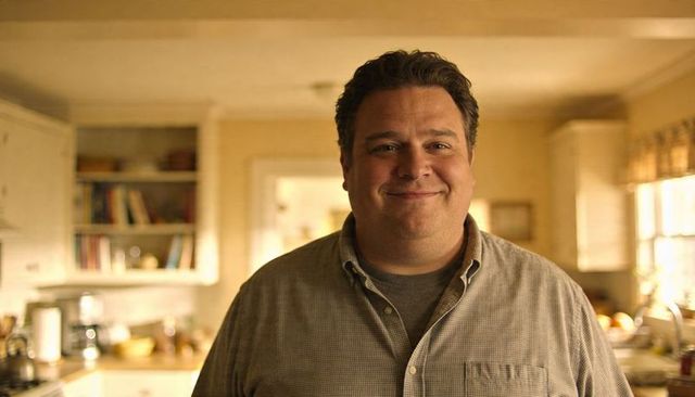 Smiling man standing in sunlit kitchen wearing button-down over gray t-shirt, warm portrait