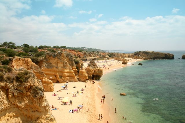 Beautiful beach scene with clear ocean water and golden sandy shore lined with rocky cliffs. Tourists are sunbathing and swimming, enjoying summer vacation activities. This picturesque landscape is perfect for promoting travel destinations, summer holidays, and coastal tourism. It can be used in travel brochures, holiday websites, and vacation planning materials.